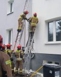 Dwóch strażaków na drabinach przy domu jednorodzinnym, za nimi grupa strażaków