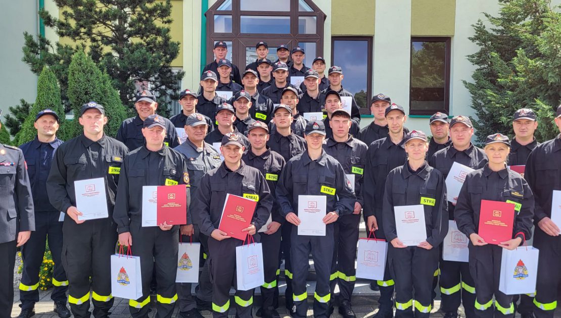 Grupa strażaków pozuje do zdjęcia na zakończenie kursu podstawowego, w dłoniach dyplomy, po lewej kierownik kursu