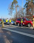 Na drodze stoją dwa uszkodzone pojazdy, dwa radiowozy oraz samochód strażacki. 4 strażaków i 3 policjantów pracują przy pojazdach.