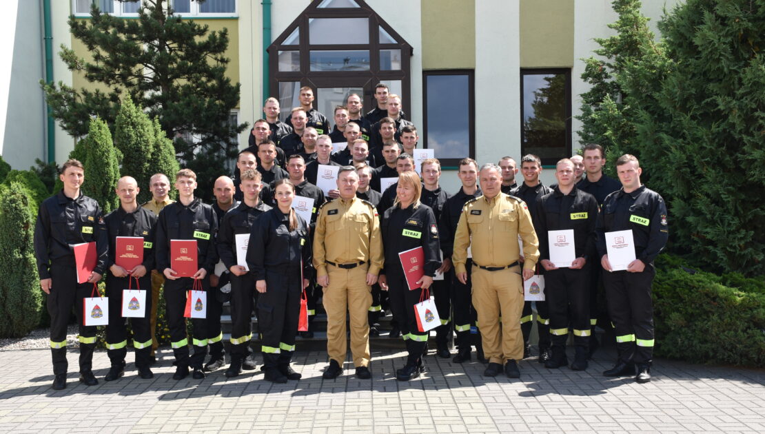 38 strażaków w mundurach koszarowych i służbowych stają , część z nich trzyma dokumenty w ręku. W tle budynek i drzewa.