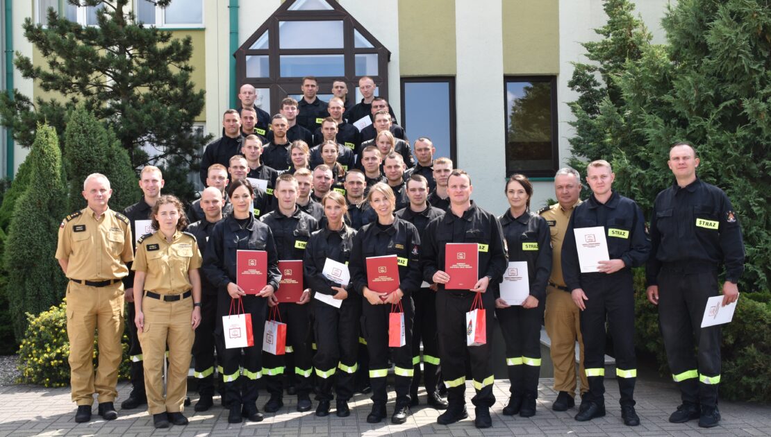 39 strażaków w mundurach koszarowych i służbowych stają , część z nich trzyma dokumenty w ręku. W tle budynek i drzewa.