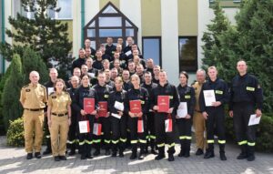 39 strażaków w mundurach koszarowych i służbowych stają , część z nich trzyma dokumenty w ręku. W tle budynek i drzewa.