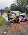 Wokół rozbitego samochodu na jezdni pracuje 3 strażaków w ubraniach specjalnych. W tle samochód strażacki i dwóch strażaków w ubraniu specjalnym.