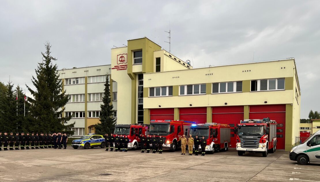 Przed budynkiem w szeregu stoi grupa strażaków. W tle stoją samochody strażackie i samochód policji