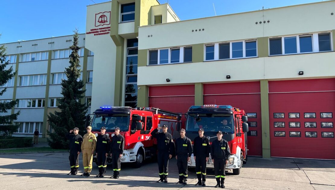 8 strażaków w mundurze koszarowym stoją w szeregu. W tle dwa wozy strażackie i budynek.