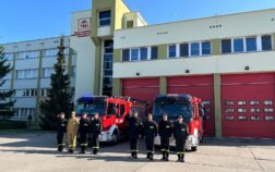 8 strażaków w mundurze koszarowym stoją w szeregu. W tle dwa wozy strażackie i budynek.