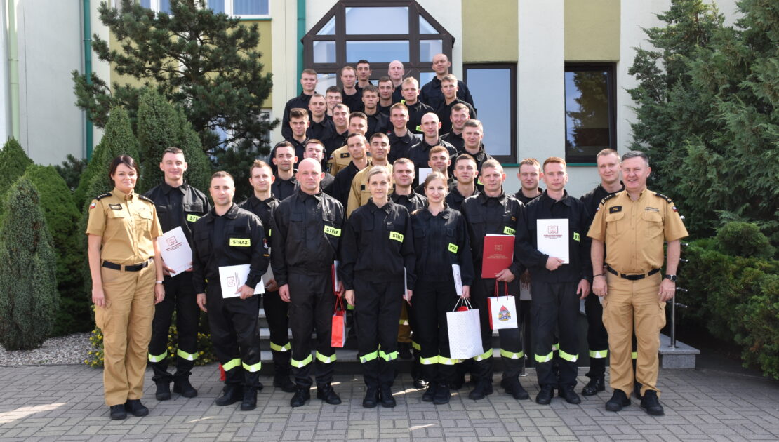 37 strażaków w mundurach koszarowych i służbowych stają , część z nich trzyma dokumenty w ręku. W tle budynek i drzewa.