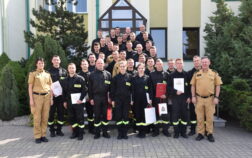 37 strażaków w mundurach koszarowych i służbowych stają , część z nich trzyma dokumenty w ręku. W tle budynek i drzewa.