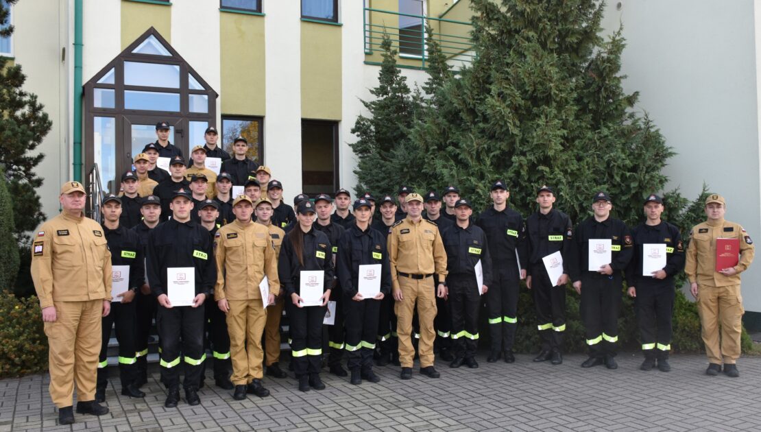 38 strażaków w mundurach koszarowych i służbowych stają , część z nich trzyma dokumenty w ręku. W tle budynek i drzewa.