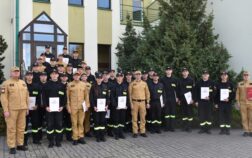 38 strażaków w mundurach koszarowych i służbowych stają , część z nich trzyma dokumenty w ręku. W tle budynek i drzewa.