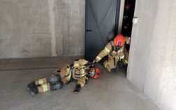 W budynku na ziemie leży strażak, który jest ciągnięty po podłodze przez drugiego strażaka. Ratownicy są w ubraniu specjalnym