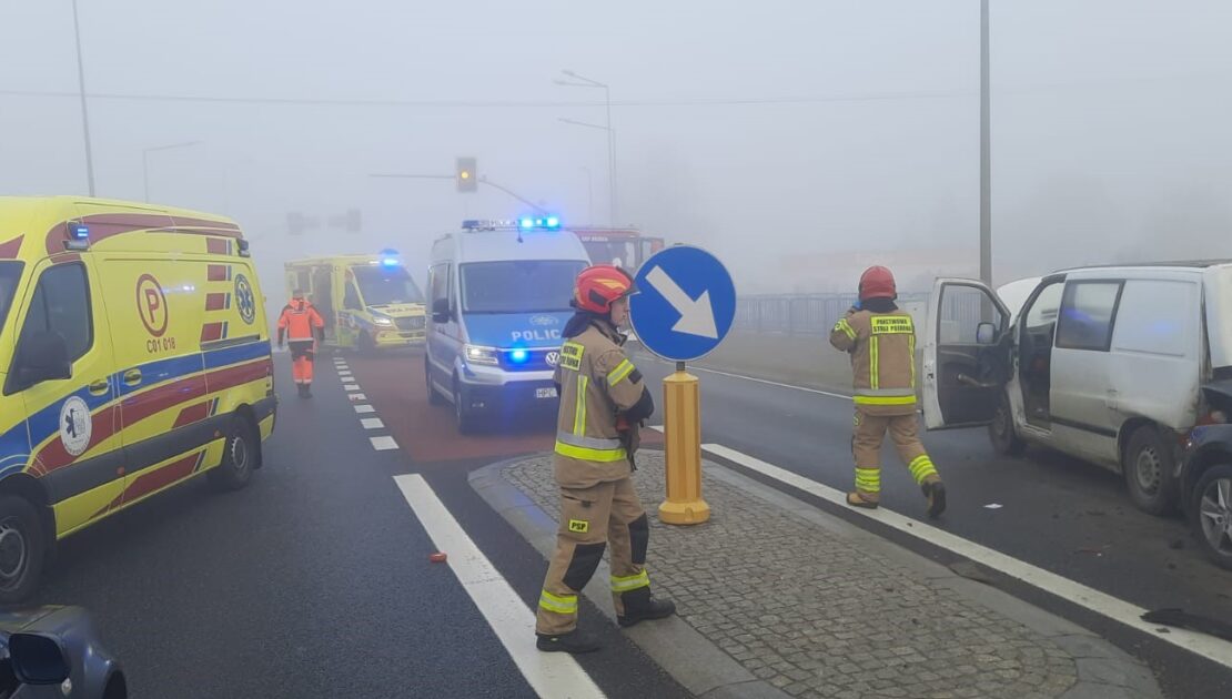 Na jezdni stoi patrol Policji i Ambulans. W tle dwóch strażaków i ratownik medyczny.