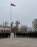 Strażacy w ubraniach koszarowych stoją na zbiórce. Na maszcie powiewa flaga Polski.