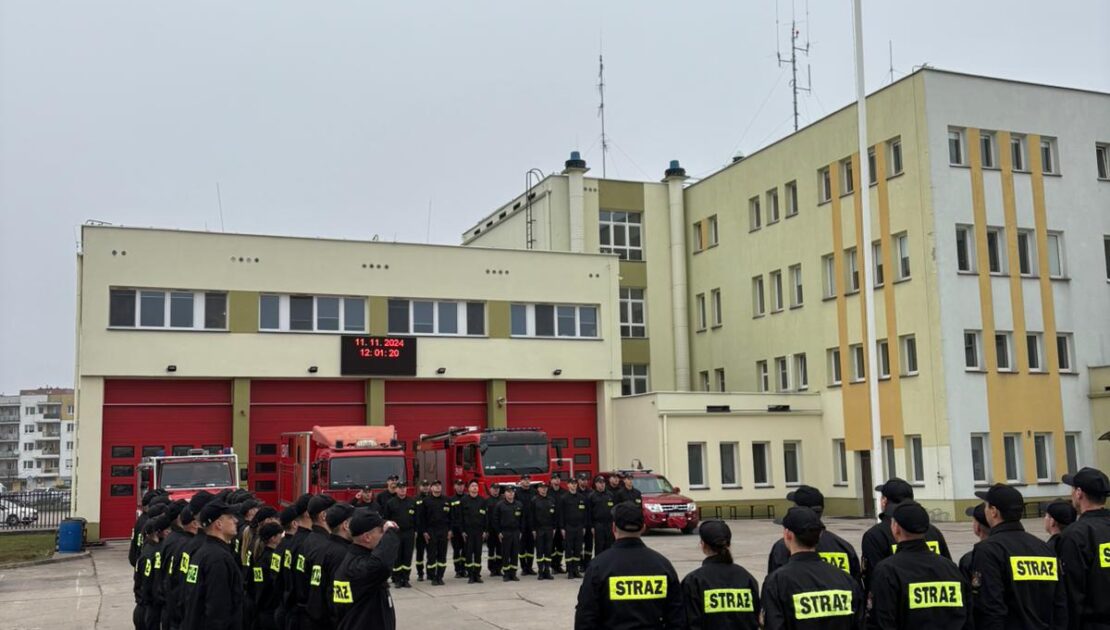 Strażacy w ubraniach koszarowych stoją na zbiórce. Na maszcie powiewa flaga Polski