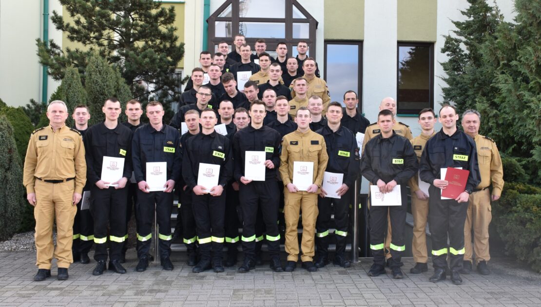 39 strażaków w mundurach koszarowych i służbowych stają , część z nich trzyma dokumenty w ręku. W tle budynek i drzewa.