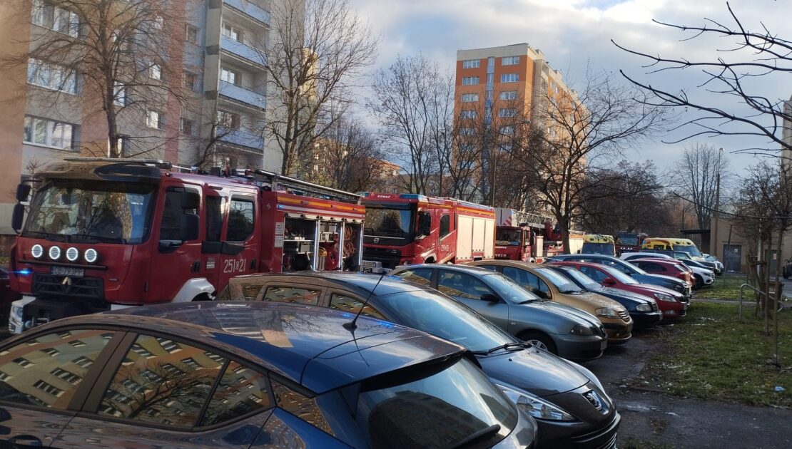 Zaparkowane samochody osobowe wzdłuż ulicy. Na ulicy stoją dwa samochody strażackie. W tle dwa bloki mieszklane.