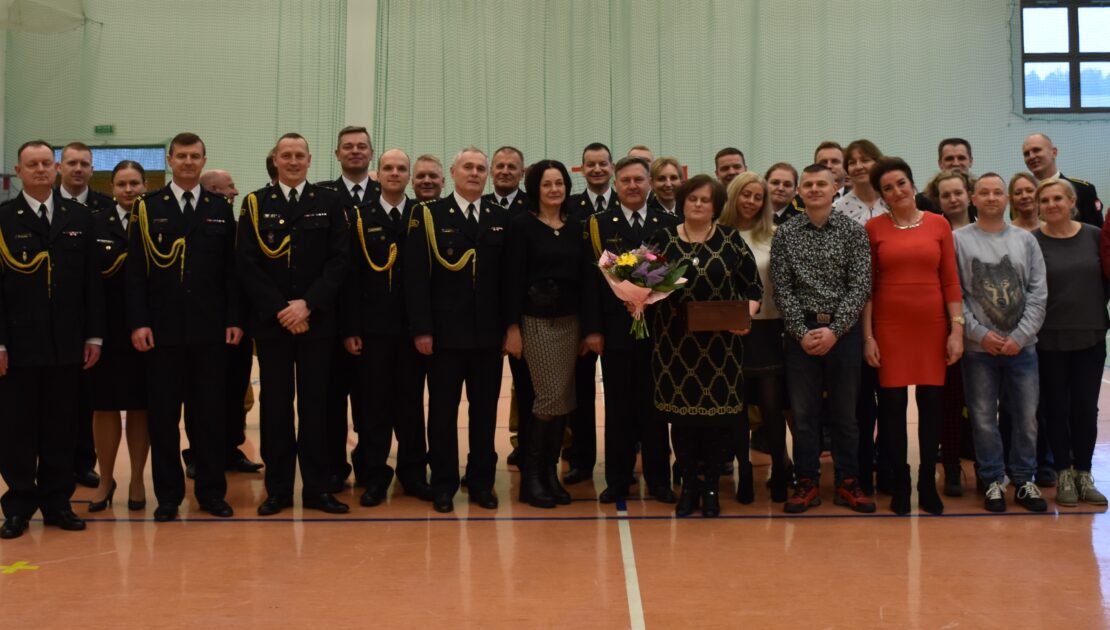 Kilkudziesięciu strażaków i osób cywilnych stoją w mundurach wyjściowych w pomieszczeniu. Jedna osoba trzyma w ręku kwiaty.