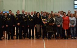 Kilkudziesięciu strażaków i osób cywilnych stoją w mundurach wyjściowych w pomieszczeniu. Jedna osoba trzyma w ręku kwiaty.